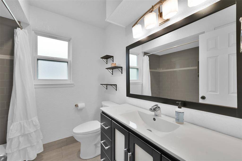 The remodeled Secondary Bathroom has a vanity with drawers and cabinet for storage, a framed mirror and updated light fixture. To the right just out of the picture is the modern tub/shower with decorative tile and product niche. There is also an additional built-in cabinet with laundry hamper tucked away behind the door.