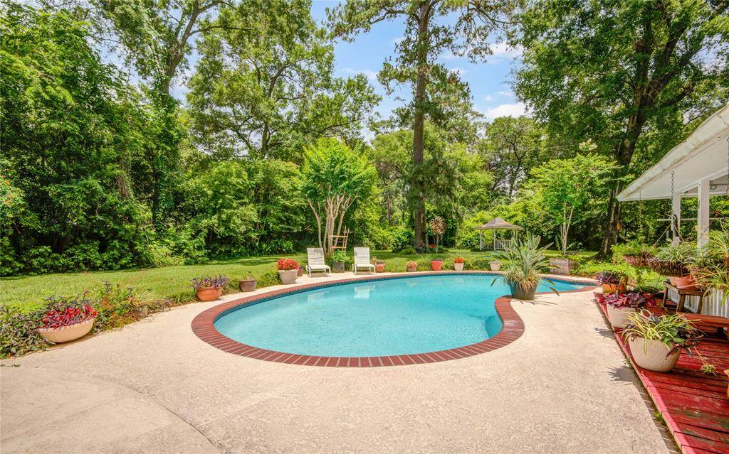 This angle highlights the sparkling pool nestled within an expansive, lushly green yard surrounded by mature trees, with a charming gazebo tucked into the rear and a generous covered entertaining space visible to the right—an idyllic outdoor retreat.