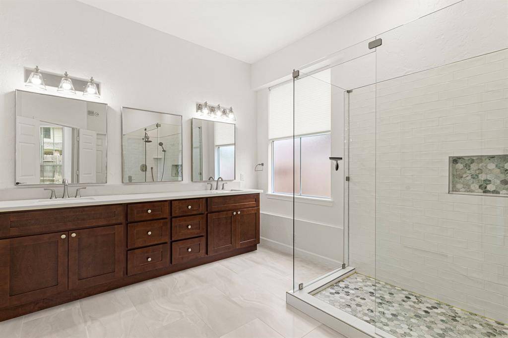 The remodeled primary bathroom has a new double vanity, mirrors, light fixtures, tile, and oversized shower with frameless glass enclosure and designer tile