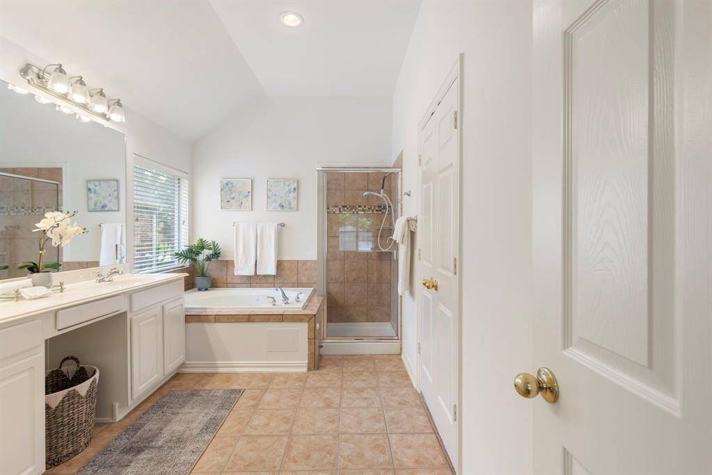 The bathroom features a spacious layout with a large vanity and mirror, tiled flooring, a soaking tub, and a separate glass-enclosed shower. Natural light streams through a window, enhancing the bright, airy feel.