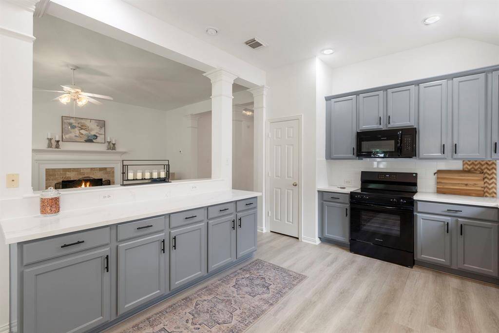 This inviting kitchen features modern gray cabinetry, sleek black appliances, and ample counter space. It opens to family room with a fireplace, creating a seamless flow ideal for entertaining.