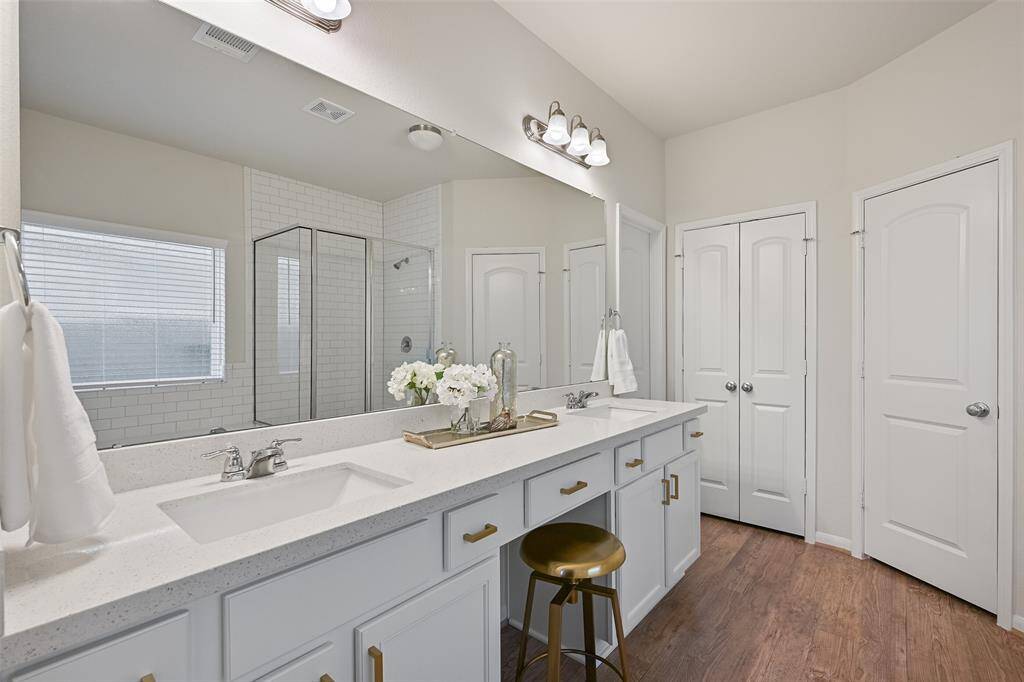 The en suite master bathroom is light and bright, offering a lengthy quartz top vanity with dual rectangular basin sinks, a seated vanity space, white cabinetry with gold hardware, luxury vinyl plank floors, and a large soaker tub with a subway tile surround that extends into the oversized glass enclosed shower.