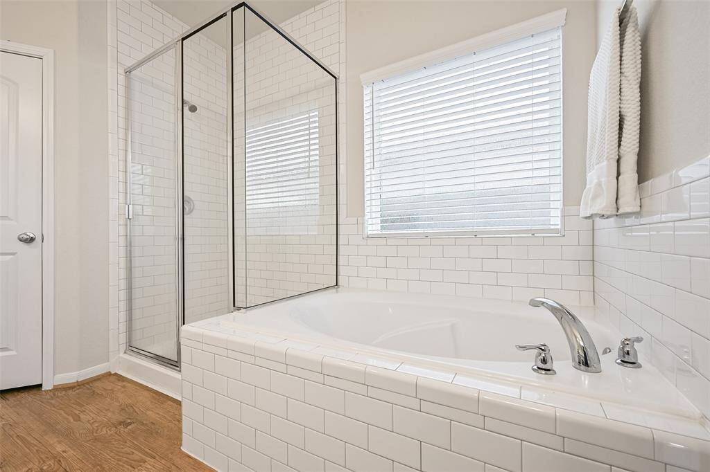The en suite master bathroom is light and bright, offering a lengthy quartz top vanity with dual rectangular basin sinks, a seated vanity space, white cabinetry with gold hardware, luxury vinyl plank floors, and a large soaker tub with a subway tile surround that extends into the oversized glass enclosed shower.
