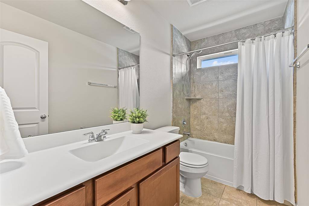 This bathroom services the gameroom and two previous bedrooms, featuring a lengthy cultured marble countertop with a rectangular basin sink, stained wood cabinetry, tile floors, and a combination bathtub and shower with a tile surround.
