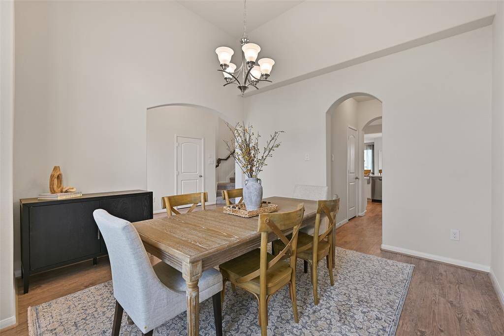 To the right of the foyer, a wide arched entrance leads into this elegant dining room with an elevated 13-foot high ceiling, continued luxury vinyl plank floors, contemporary lighting, and a pair of front-facing windows dressed with 2-inch blinds and neutral beige draperies.
