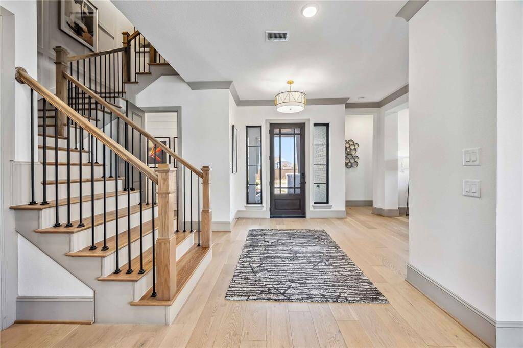 Step into a bright and welcoming entryway featuring light wood flooring, a stylish iron staircase, and double vertical windows flanking the glass-paneled front door for an elegant first impression.