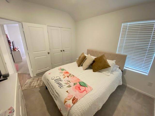 This is a bright and neatly presented bedroom featuring a large window with blinds, a plush bed with decorative pillows, a double-door closet, and tiled flooring. It has a neutral color scheme and connects to another room.
