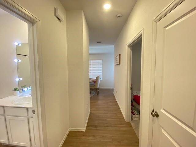 This is a bright, well-lit hallway with modern wooden flooring, leading to a room with a window. On the left, there's a bathroom vanity with a stylish mirror and lighting, indicating a bathroom entrance. The space appears clean and well-maintained.