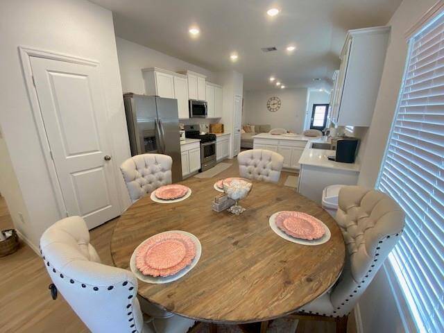 This image showcases a modern, open-plan dining and kitchen area with contemporary finishes, featuring a round wood dining table with plush, tufted chairs, stainless steel appliances, white cabinetry, and recessed lighting. The space is well-lit with natural light from large windows adorned with blinds.