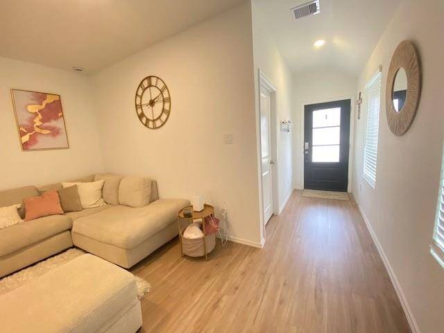 This is a bright, modern living space featuring a neutral color palette, wood-like flooring, and contemporary decor. The room has natural light coming from windows near the front door, enhancing the welcoming atmosphere.
