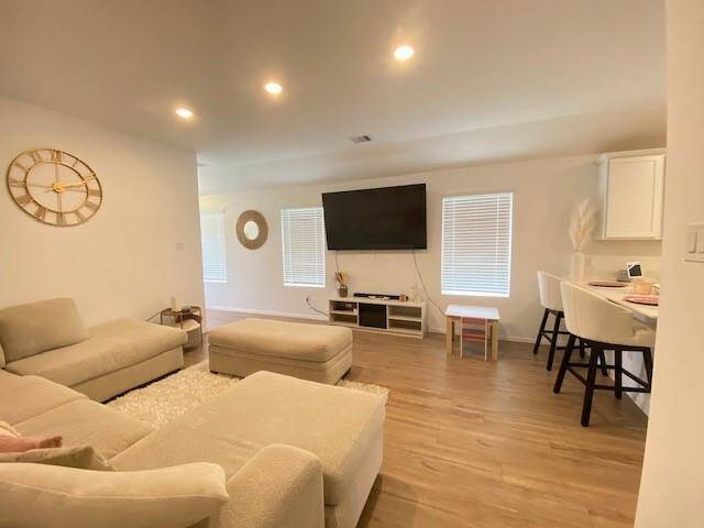 The photo shows a modern, open-plan living space with recessed lighting, hardwood floors, and neutral colors. The area is furnished with a comfortable sectional sofa, a large flat-screen TV, and a dining area with a bar-style table and chairs. The room feels bright and welcoming with natural light coming through the two windows with blinds.