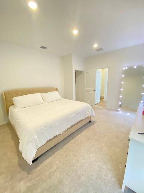This is a bright, spacious bedroom with carpeted flooring, featuring a large bed, recessed lighting, and access to an adjoining room through a white door.