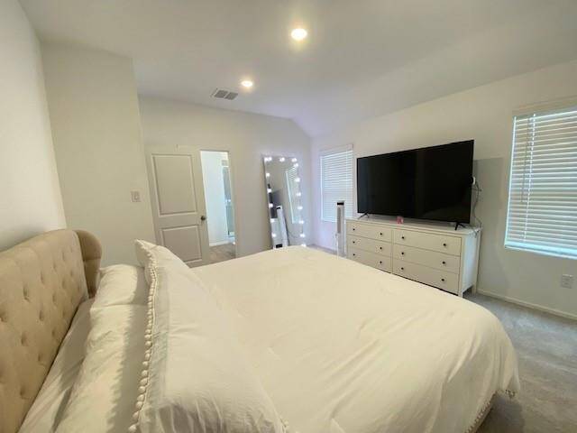 This is a modern, well-lit bedroom featuring a large bed with a tufted headboard, a dresser with a TV above, and a vanity mirror near the entrance. The room has a clean, neutral color palette and is equipped with recessed lighting and window blinds for adjustable natural light.