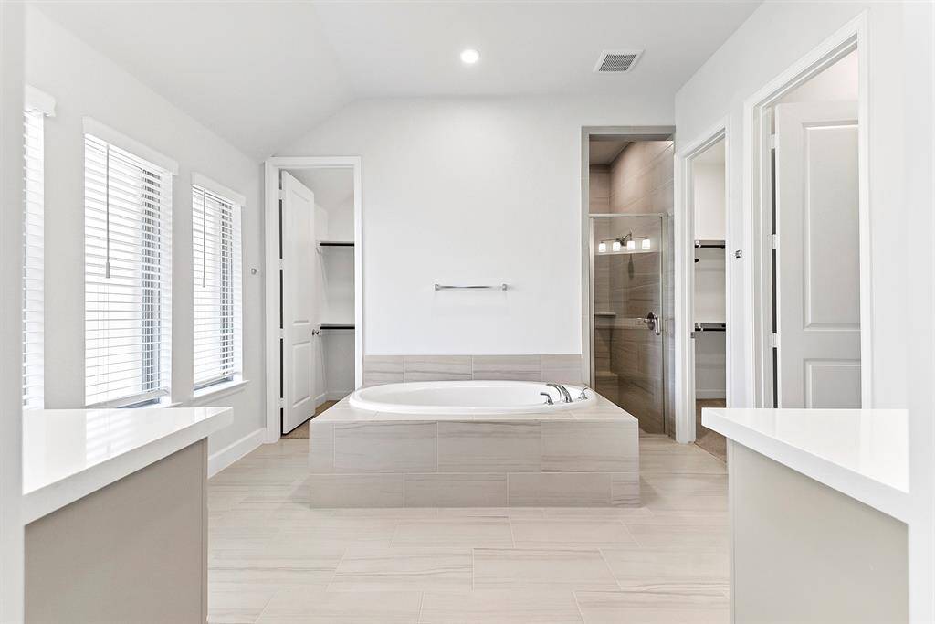 Welcome to the spa-like primary bathroom, featuring dual vanity sinks, a center island for added convenience, a soaking tub separate from the glass-enclosed tile shower, and two spacious walk-in wardrobe closets.