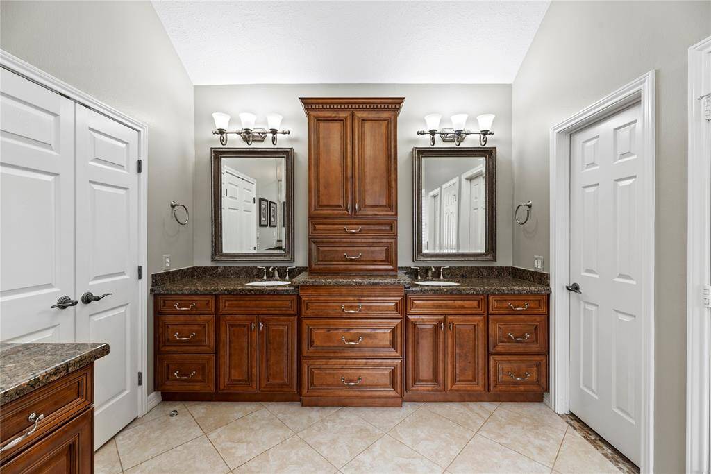 This bathroom features a double vanity with rich wood cabinetry and granite countertops. It includes ample storage space and elegant lighting fixtures above each mirror. The room is well-lit with a neutral color palette and tiled flooring.