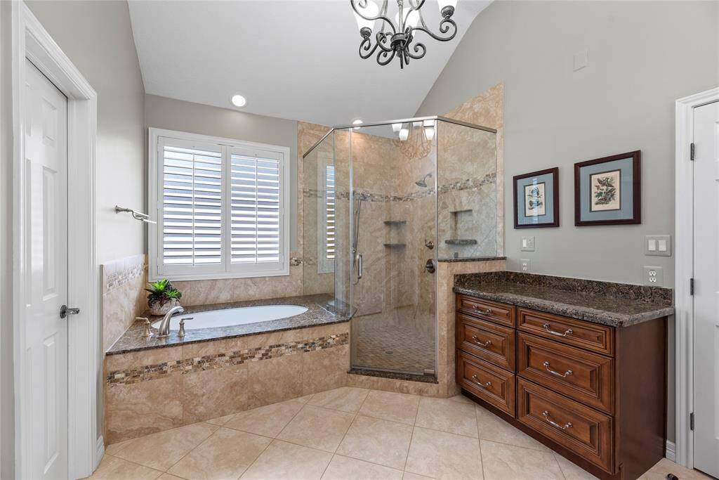 This bathroom features a luxurious design with a glass-enclosed shower, a built-in bathtub with granite surround, and elegant tile work. It includes a wooden vanity with granite countertop, modern fixtures, and natural light from the window with plantation shutters.