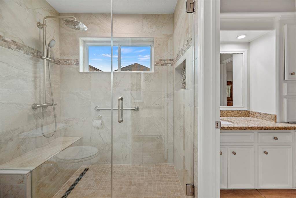 This bathroom features an updated spacious walk-in shower with elegant tile work, a rainfall showerhead, and a built-in bench. The adjoining area includes a countertop with ample cabinet storage. Natural light streams in through a window, adding brightness.
