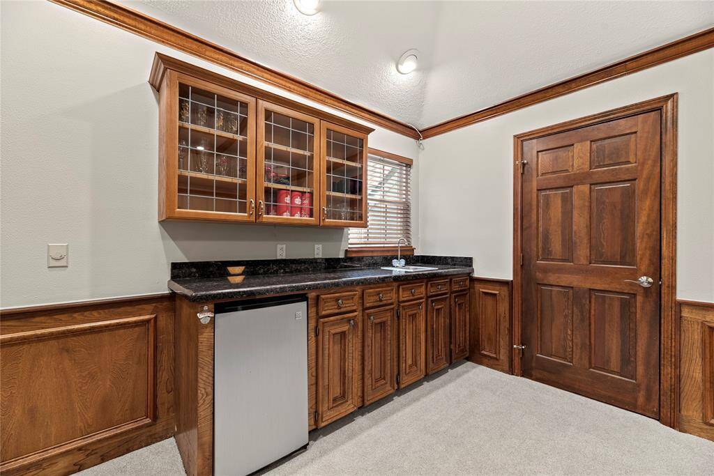 This room features a built-in wet bar with wood cabinetry, glass-front upper cabinets, and a granite countertop. It includes a small refrigerator and a sink, accented by wood paneling and crown molding for a classic touch.