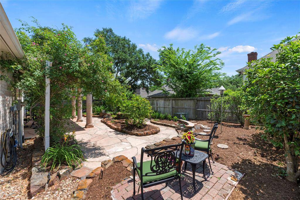 This photo showcases a charming backyard with a stone patio and lush greenery. There are seating areas with room for chairs and a small table, offering a perfect spot for relaxation. Mature trees and well-maintained plants provide a serene and private atmosphere.
