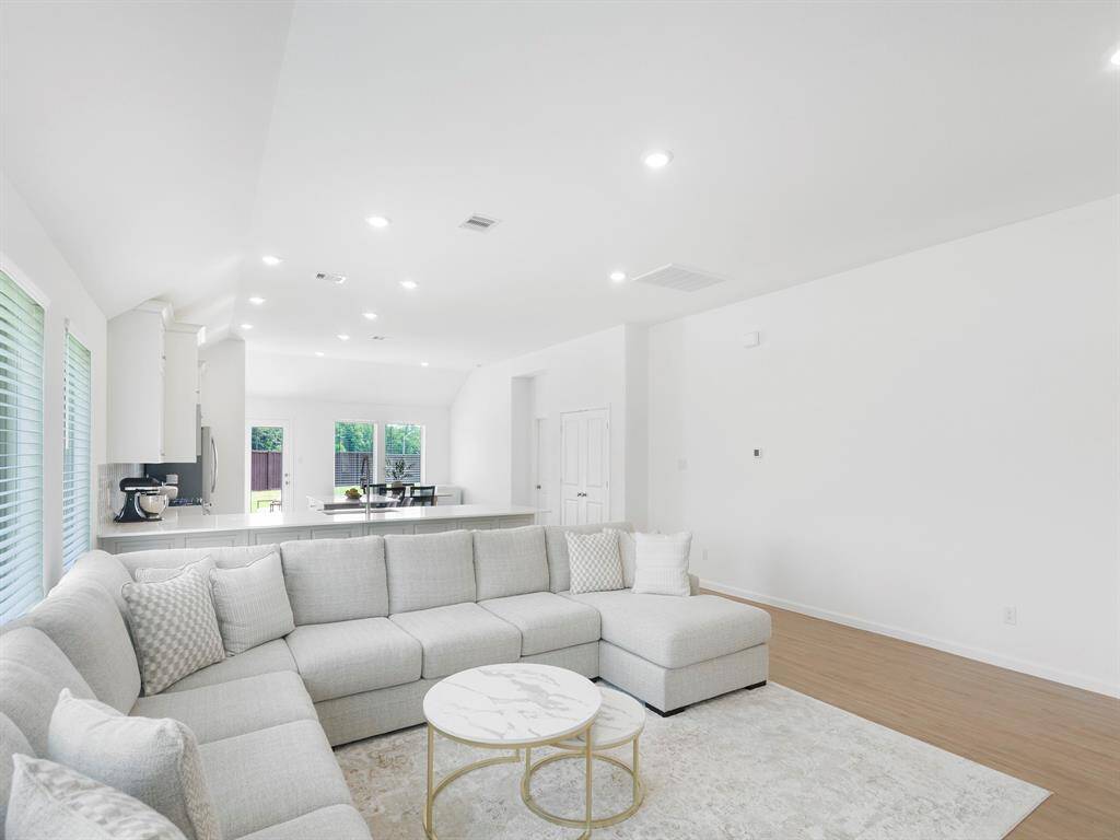 This bright, open-concept living area features a spacious L-shaped sofa, light hardwood floors, and a modern kitchen with white cabinetry. Large windows and glass doors provide ample natural light, creating an inviting and airy atmosphere.