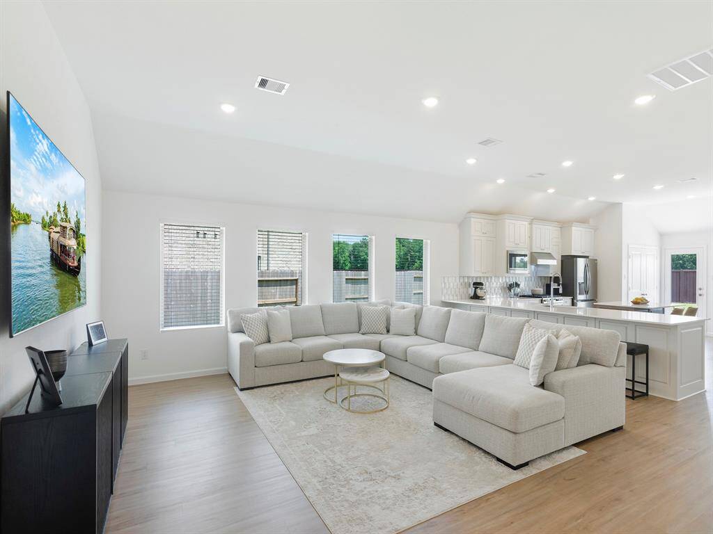 The living room features a clean, minimalist design with durable, low-maintenance flooring that mimics the look of natural wood. The wall of windows brings in ample natural light, highlighting the spaciousness created by the high ceilings. The overall style is simple and modern, offering a bright, open feel that's both functional and inviting.