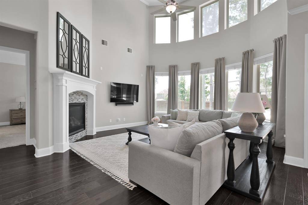 This spacious living room features high ceilings and large windows that provide ample natural light. It includes a cozy fireplace and elegant warm hardwood floors. The neutral-tones and spacious layout create a welcoming atmosphere, perfect for relaxation or entertaining.