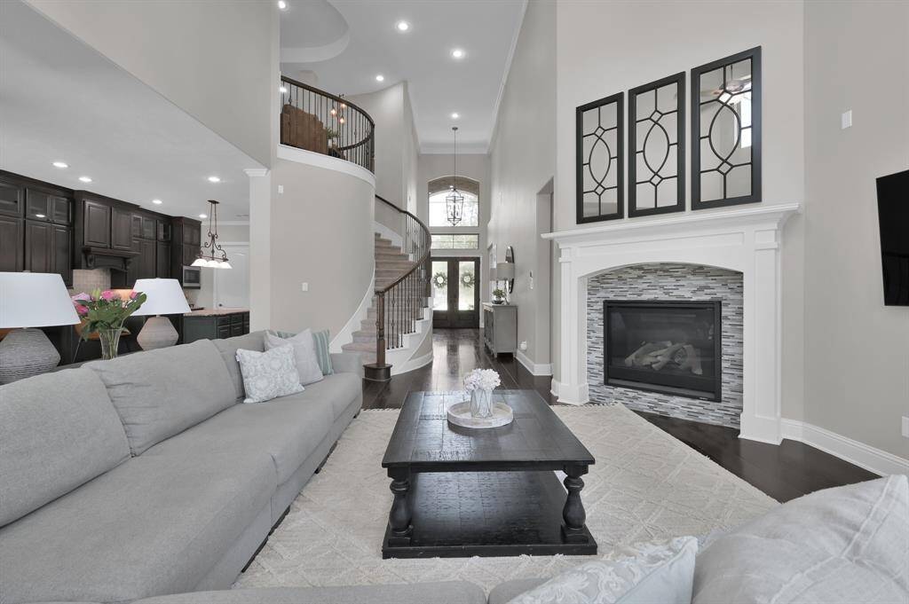 This spacious living room features a high ceiling and an elegant fireplace with modern tile accents. The open-concept design flows into a sleek kitchen with dark cabinetry. A grand staircase leads to the upper level, enhancing the room's sophisticated ambiance.