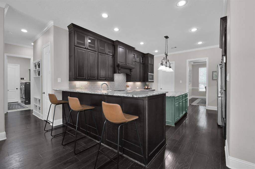 This kitchen features a modern design with dark wood cabinets, a large island with granite countertops, and stylish pendant lighting. There is a spacious breakfast bar, stainless steel appliances, and ample natural light. The space connects seamlessly to other parts of the home, offering an open and inviting atmosphere.