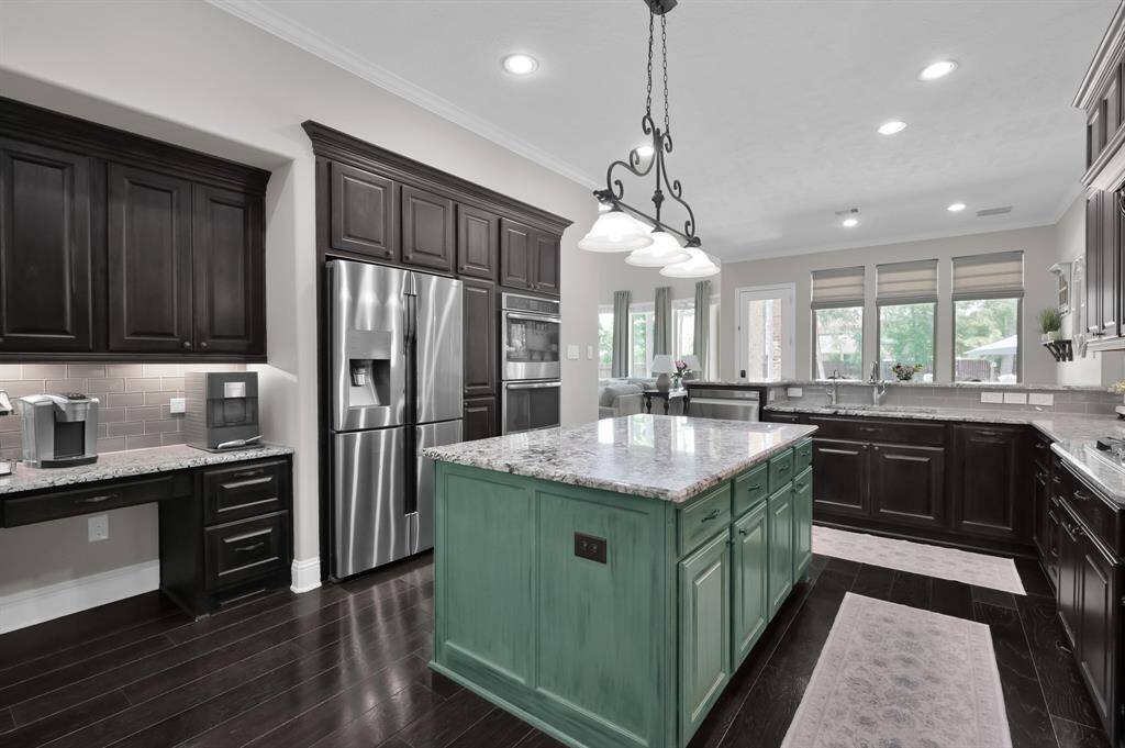 This kitchen features dark wood cabinetry, a large central island with a granite countertop, and stainless steel appliances. It includes ample natural light from multiple windows and has a cozy built-in desk area. The lighting fixtures and dark wood floors add elegance and warmth to the space.