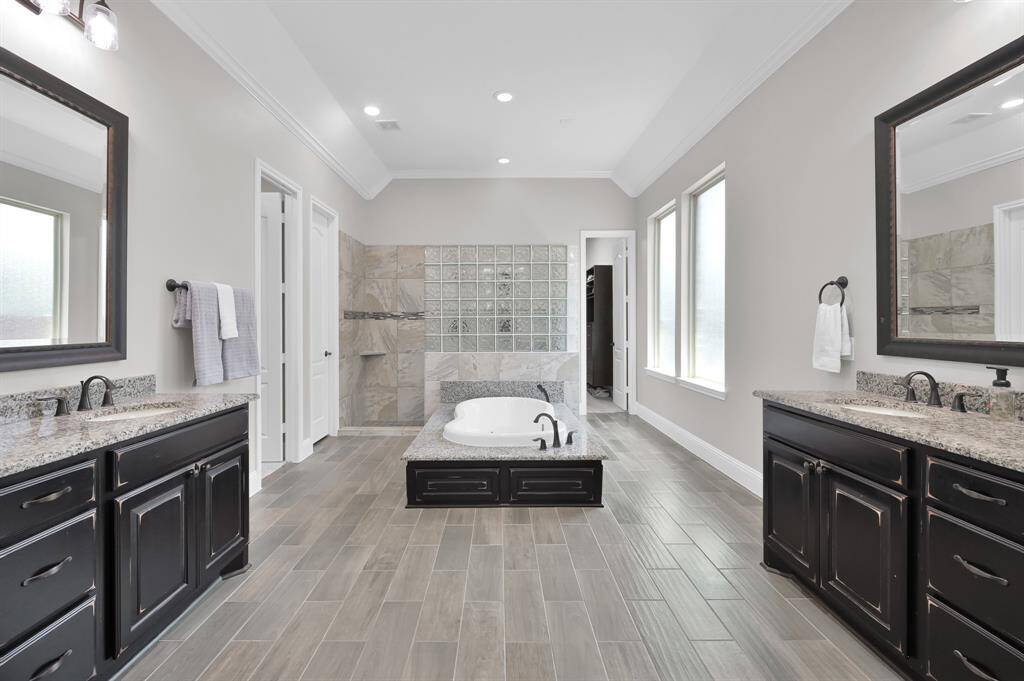 This spacious bathroom features dual vanities with granite countertops, a central soaking tub, and a walk-in shower. The room is enhanced by elegant tile flooring, large mirrors, and ample natural light from the windows.
