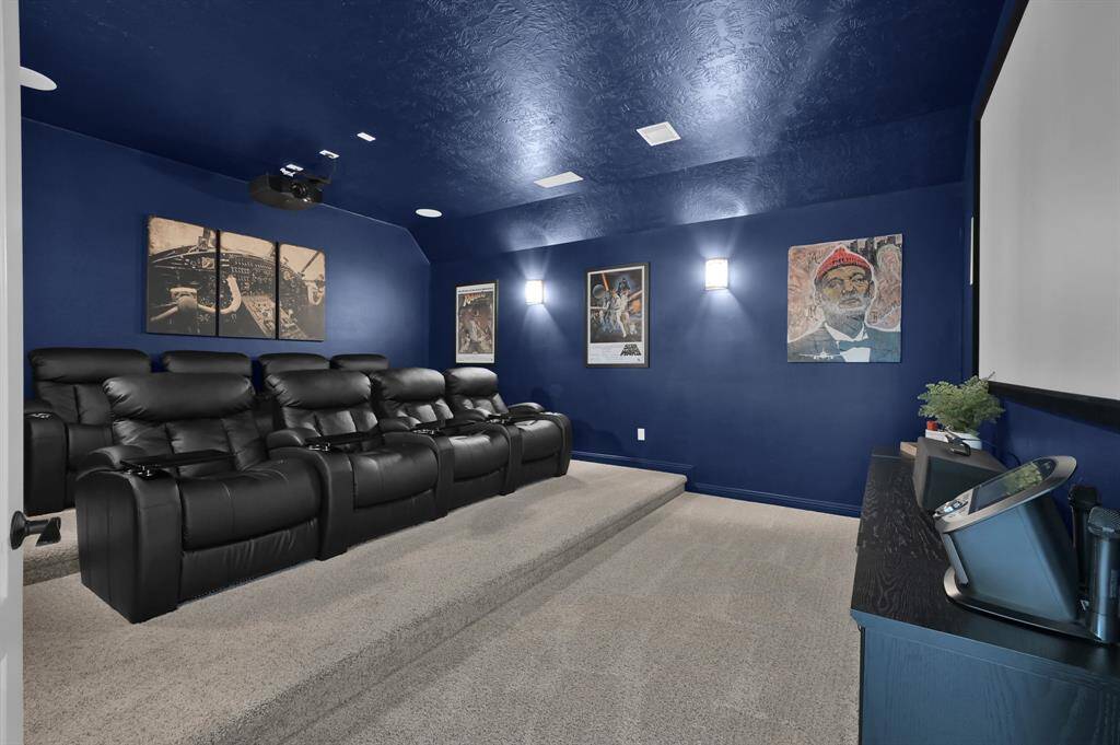 This photo showcases a spacious but cozy home theater with dark blue walls, featuring two rows of comfortable black recliners (8 total) on a carpeted double platform. The room's seating, movie equipment and massive 120" screen, will remain with the home.