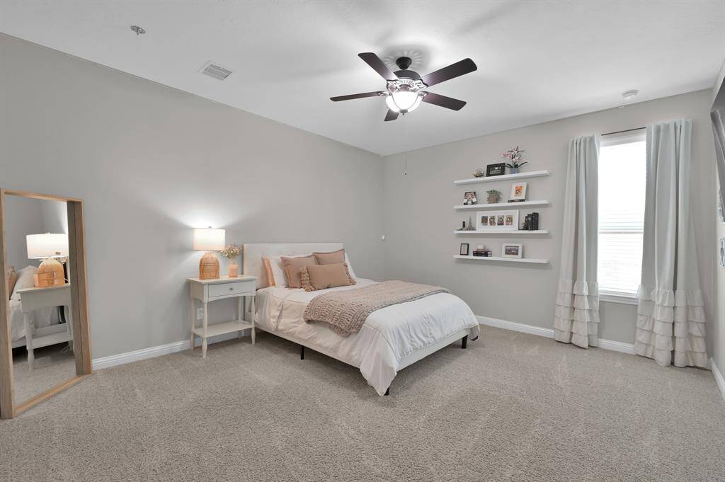 This bedroom is spacious and inviting, featuring neutral tones and carpeted flooring. It includes a ceiling fan, a large window for natural light, and plenty of space to spread out and personalize the space.