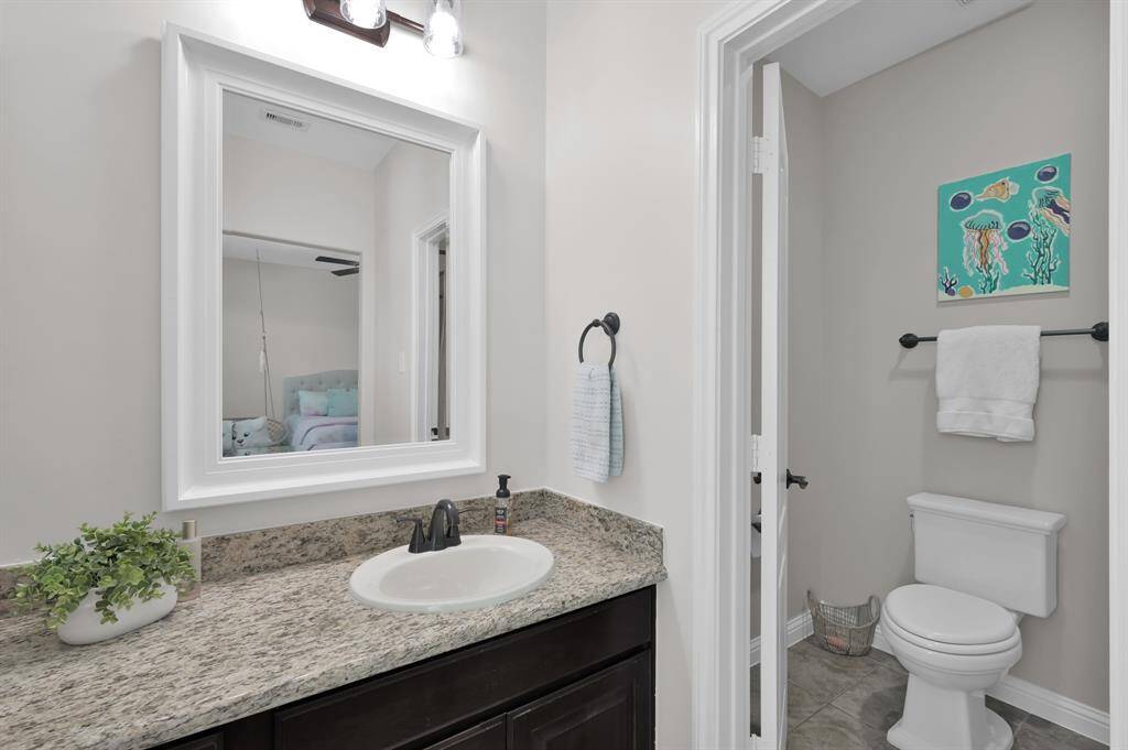 This photo shows a bright, modern bathroom. It features a granite countertop with a single sink, framed mirror, and stylish light fixtures. The space includes a separate toilet area that includes a tub-shower and an adjoining second sink area.  This is one half of the Jack and Jill bathroom.