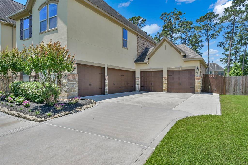 This photo showcases a modern home exterior with a spacious driveway leading to five-car garages. The property features well-maintained landscaping with flowering plants and trees, and is set against a backdrop of tall trees, offering a blend of nature and suburban comfort.