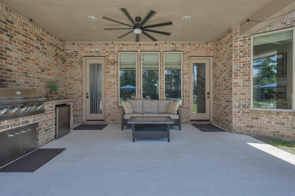 Spacious covered patio with brick walls, featuring a built-in stainless steel grill and mini fridge. Equipped with a ceiling fan, it has a cozy seating area and plenty of space for outdoor entertaining.
