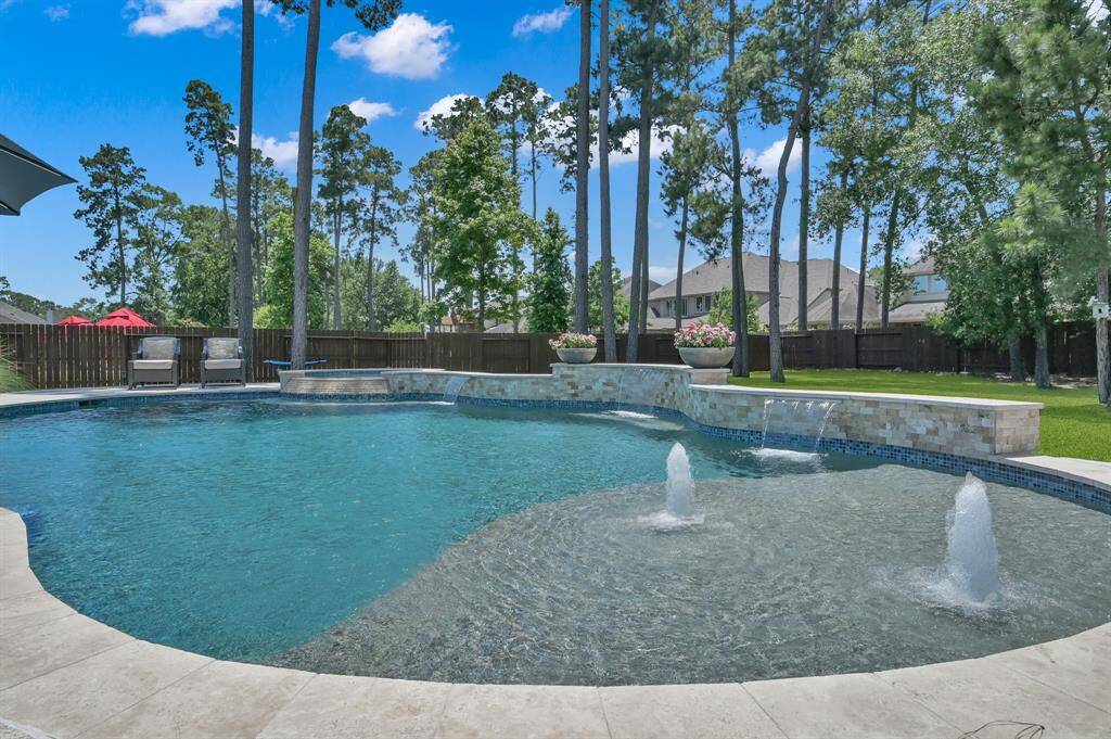 This photo showcases a spacious backyard with a luxurious pool featuring fountains and a waterfall. Surrounded by tall trees and a privacy fence, the area offers a serene and private setting, perfect for relaxation and entertaining.