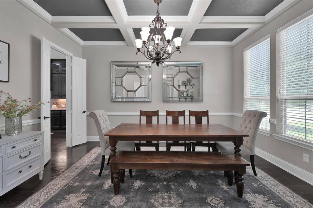This elegant dining room features a coffered ceiling with a stylish chandelier, large windows for natural light. The neutral color palette adds a touch of sophistication.