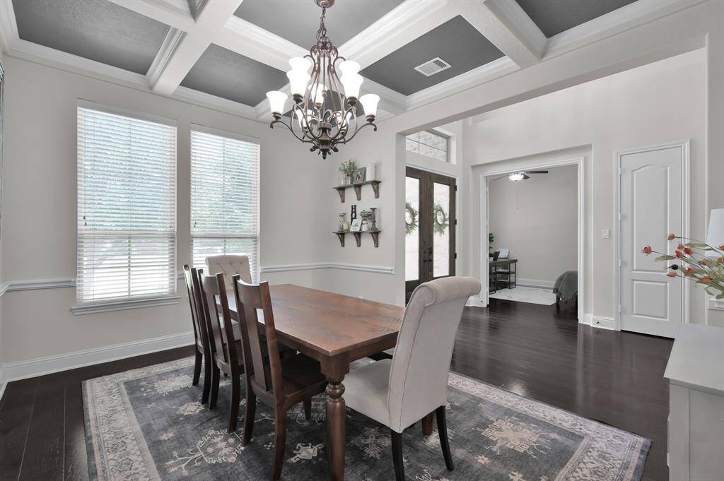 This dining room features elegant warm wood flooring, a coffered ceiling with a stylish chandelier, and abundant natural light from large windows. The space is open and inviting, with a view into the entry and adjacent rooms.