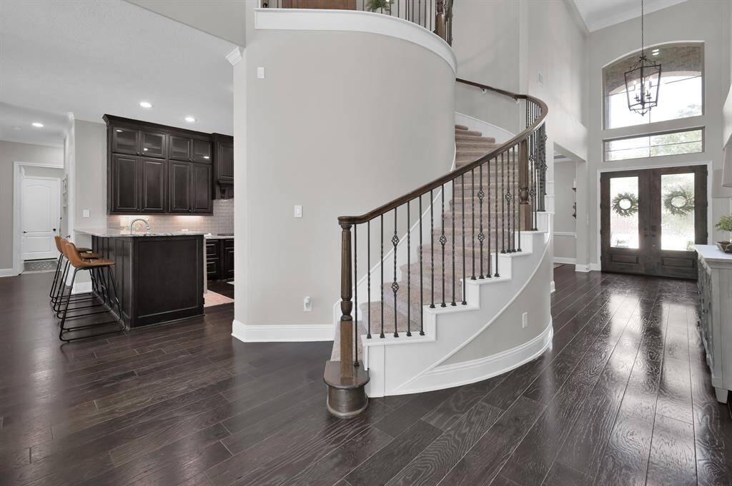 This photo showcases a stylish entryway with a grand curved staircase and warm hardwood floors. The area is bright and open, featuring double front doors with decorative glass and a glimpse into a modern kitchen with dark cabinetry and bar seating.