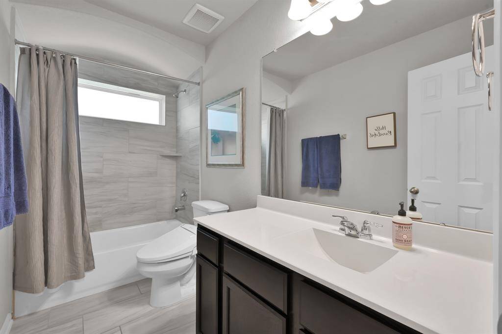 This spacious bathroom features a modern design with light gray tile floors and walls. It includes a bathtub with a shower, a large mirror above a sleek vanity with dark wood cabinets.