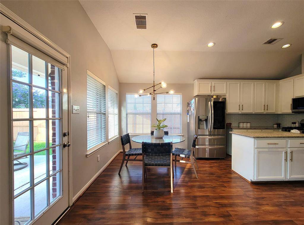 Bright and welcoming kitchen with modern appliances, white cabinetry, and warm wood floors. The dining area features a stylish light fixture and a clear view of the fenced backyard through large windows and a glass door. Ideal for family gatherings and entertaining.