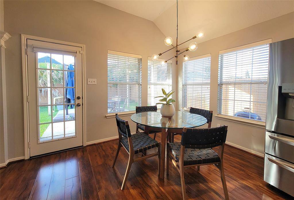 Bright dining area with hardwood floors, featuring a modern chandelier above a round glass table with four chairs. Large windows and a glass door provide ample natural light and a view of the backyard.