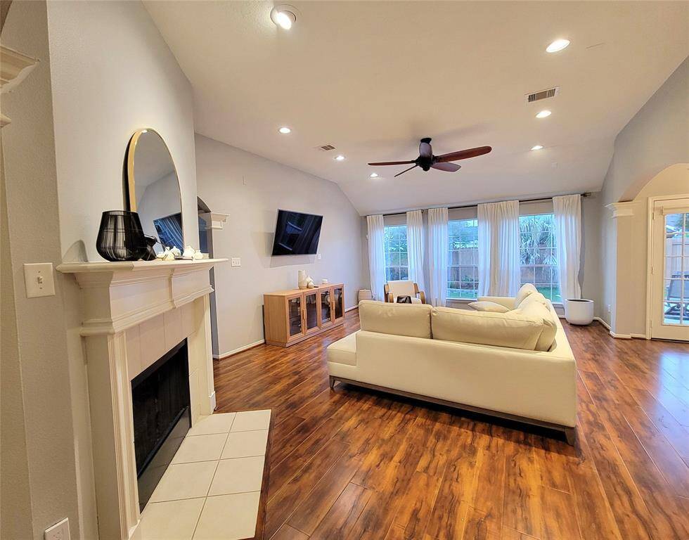 Spacious living room with warm wood flooring, a cozy fireplace, and large windows offering plenty of natural light. It features a modern ceiling fan, elegant decor, and a comfortable seating area.