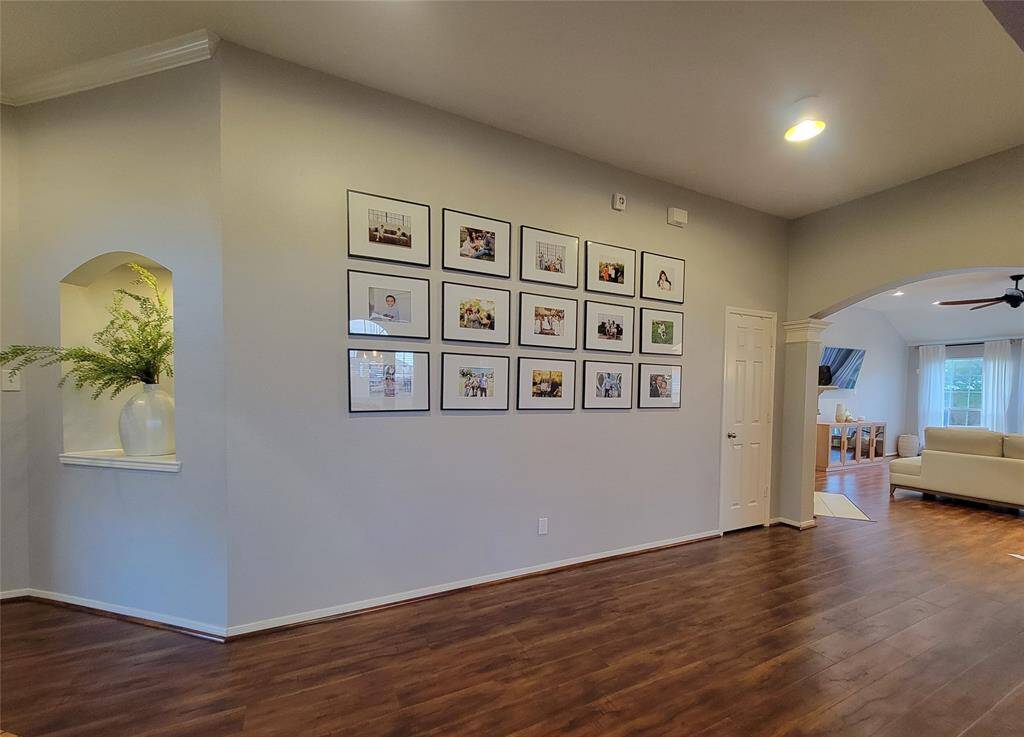 This photo showcases a spacious living area with elegant wood flooring and neutral walls. It features a gallery wall with framed photos and an arched entryway leading to a well-lit dining and living space. A decorative niche with a vase and greenery adds charm to the room.