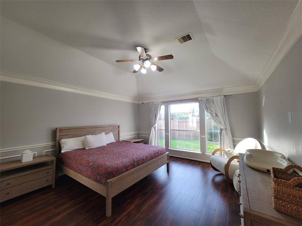 This photo shows a spacious bedroom with a vaulted ceiling and large window providing ample natural light. The room features wood flooring, a ceiling fan, and neutral-colored walls, creating a cozy and inviting atmosphere.