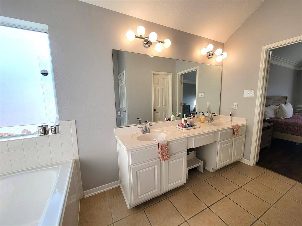 A bright, spacious bathroom with dual sinks, a large mirror, modern lighting, and a soaking tub. It connects to a bedroom, offering convenience and style.
