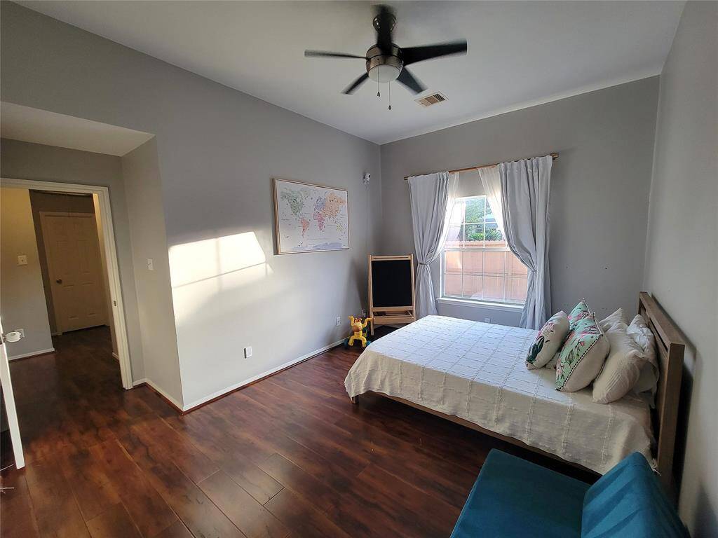 This spacious bedroom features hardwood floors, a large window with natural light, and neutral gray walls.