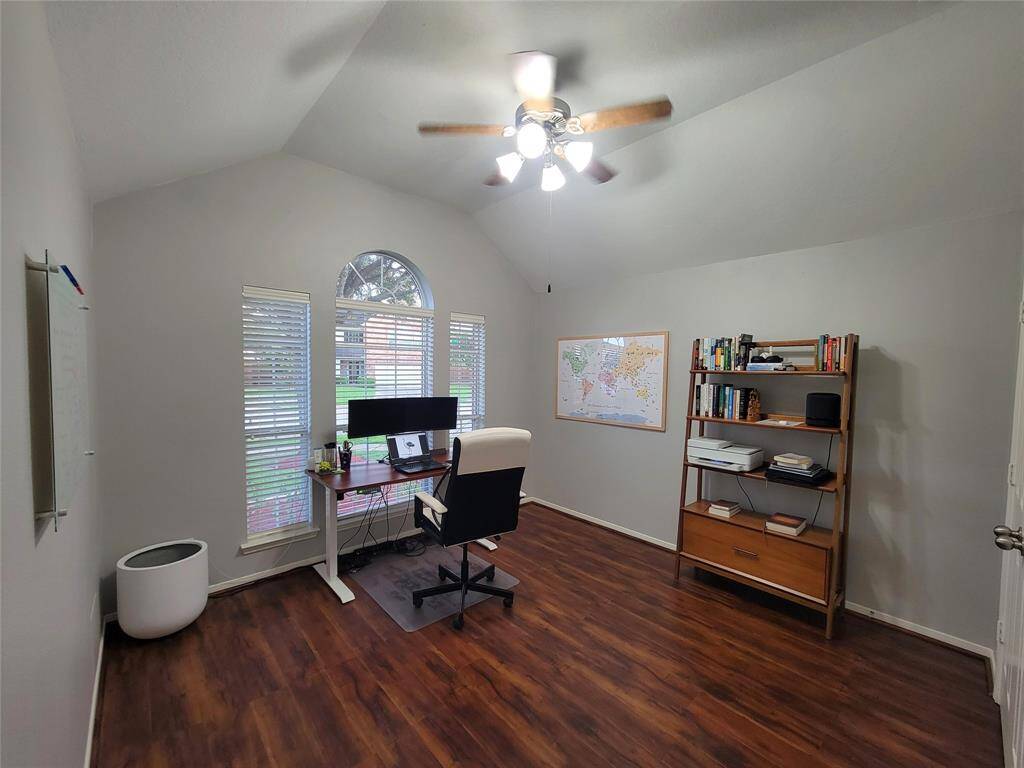 Well-lit home office with a vaulted ceiling and wood flooring. It features a large window and ceiling fan.