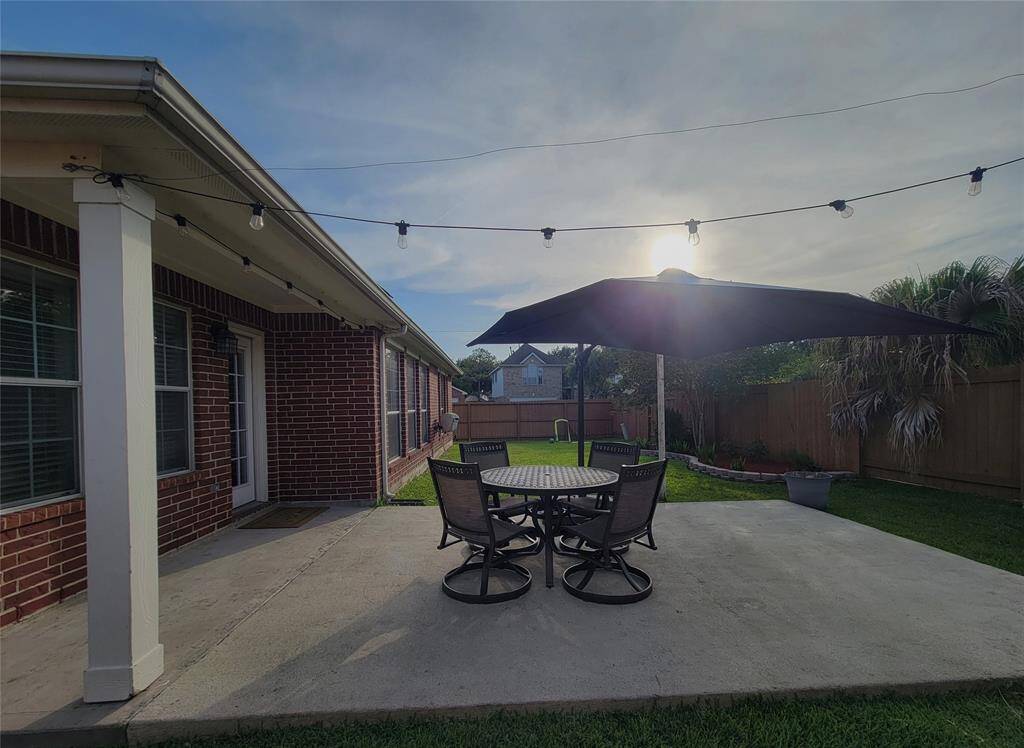 This photo showcases a cozy backyard patio with a large umbrella and seating area on a concrete slab, surrounded by a grassy lawn and a wooden fence. The brick exterior of the home adds a classic touch, and string lights provide charming ambiance. Perfect for outdoor entertaining.