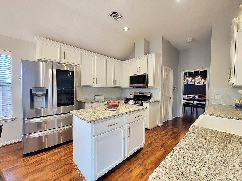 This kitchen features modern stainless steel appliances, white cabinetry, and granite countertops. It has a central island, wood flooring, and an open layout leading to a dining area with stylish lighting. Refrigerator stays!